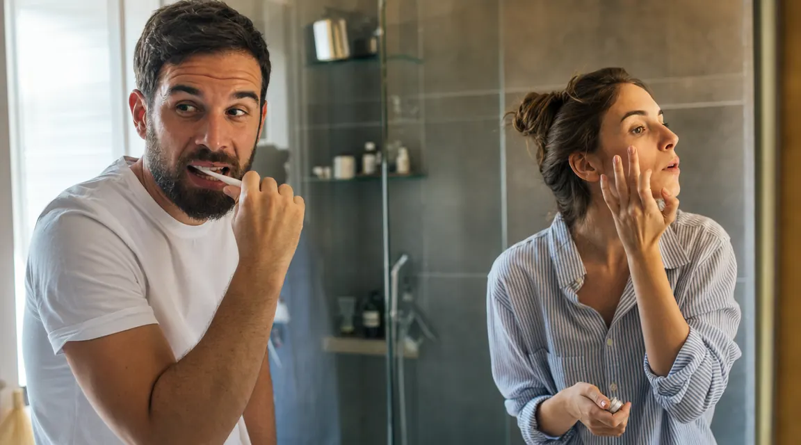 Couple in a shared bathroom, man grooming his beard while woman applies skincare nearby