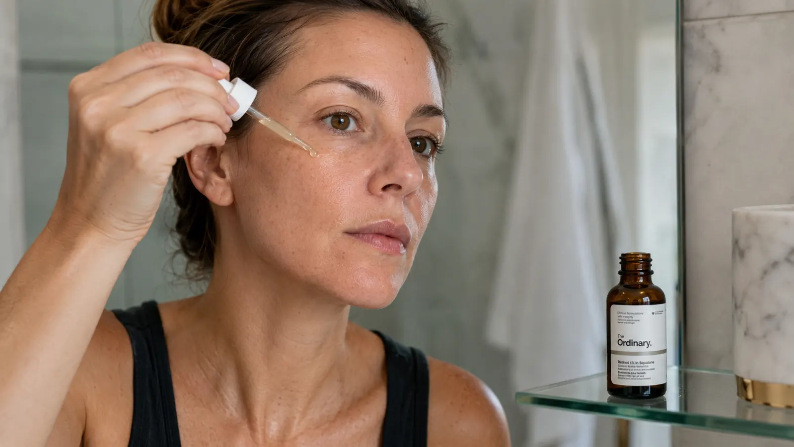 Close-up of hands applying a drop of retinol serum to a clean face in a softly lit bathroom