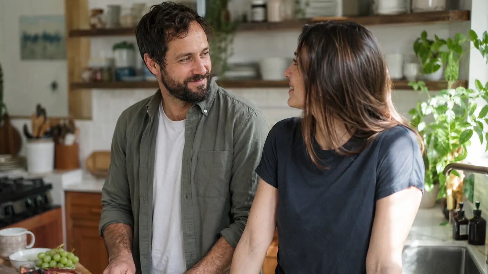 Couple in a relaxed kitchen setting, man with a groomed beard, partner looking over approvingly