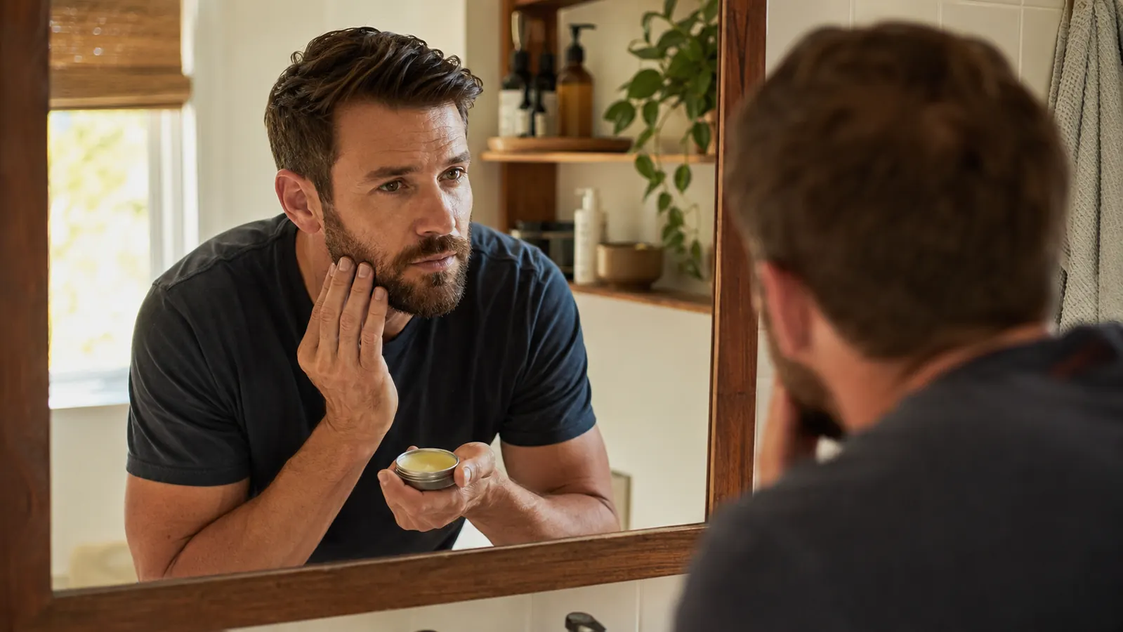 Man with a well-groomed short beard applying beard balm in a warm bathroom mirror, morning light
