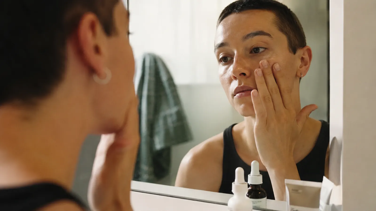 A person patting serum onto their cheekbone in front of a bathroom mirror during a morning skincare routine