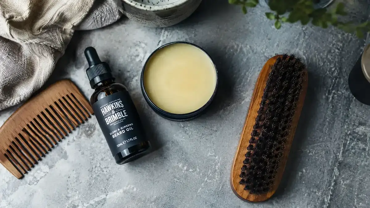 Flat-lay of beard grooming tools including oil, balm tin, wooden comb, and boar bristle brush on a stone surface
