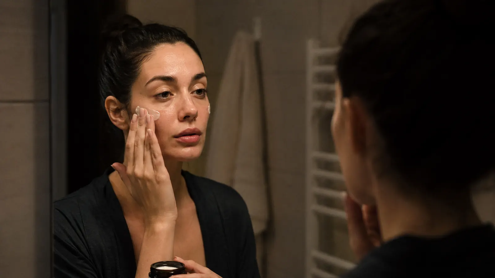 Person applying a thin layer of moisturizer over their face in a bathroom mirror, gentle technique