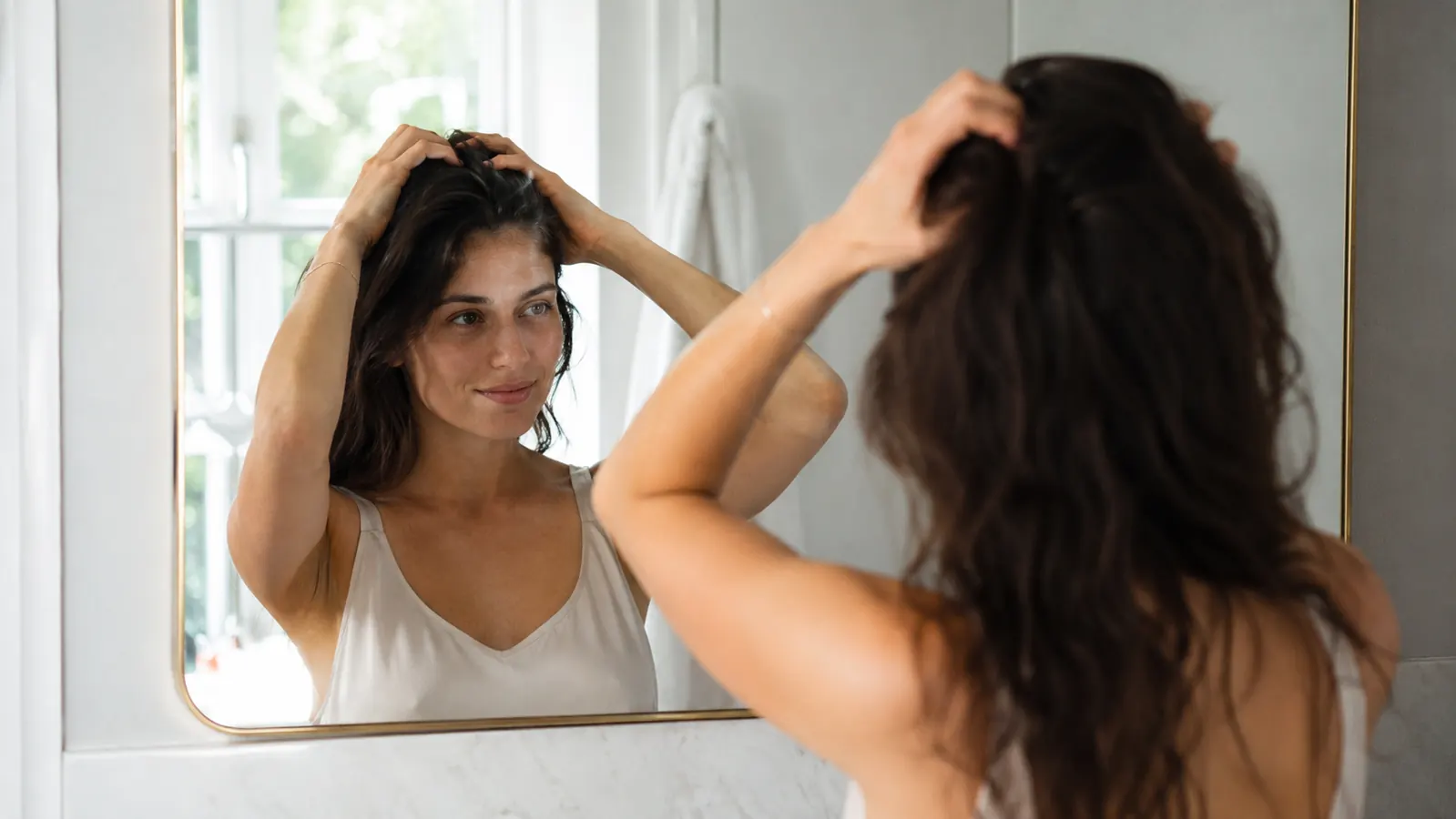 Person gently massaging scalp oil into hair roots while looking in the bathroom mirror