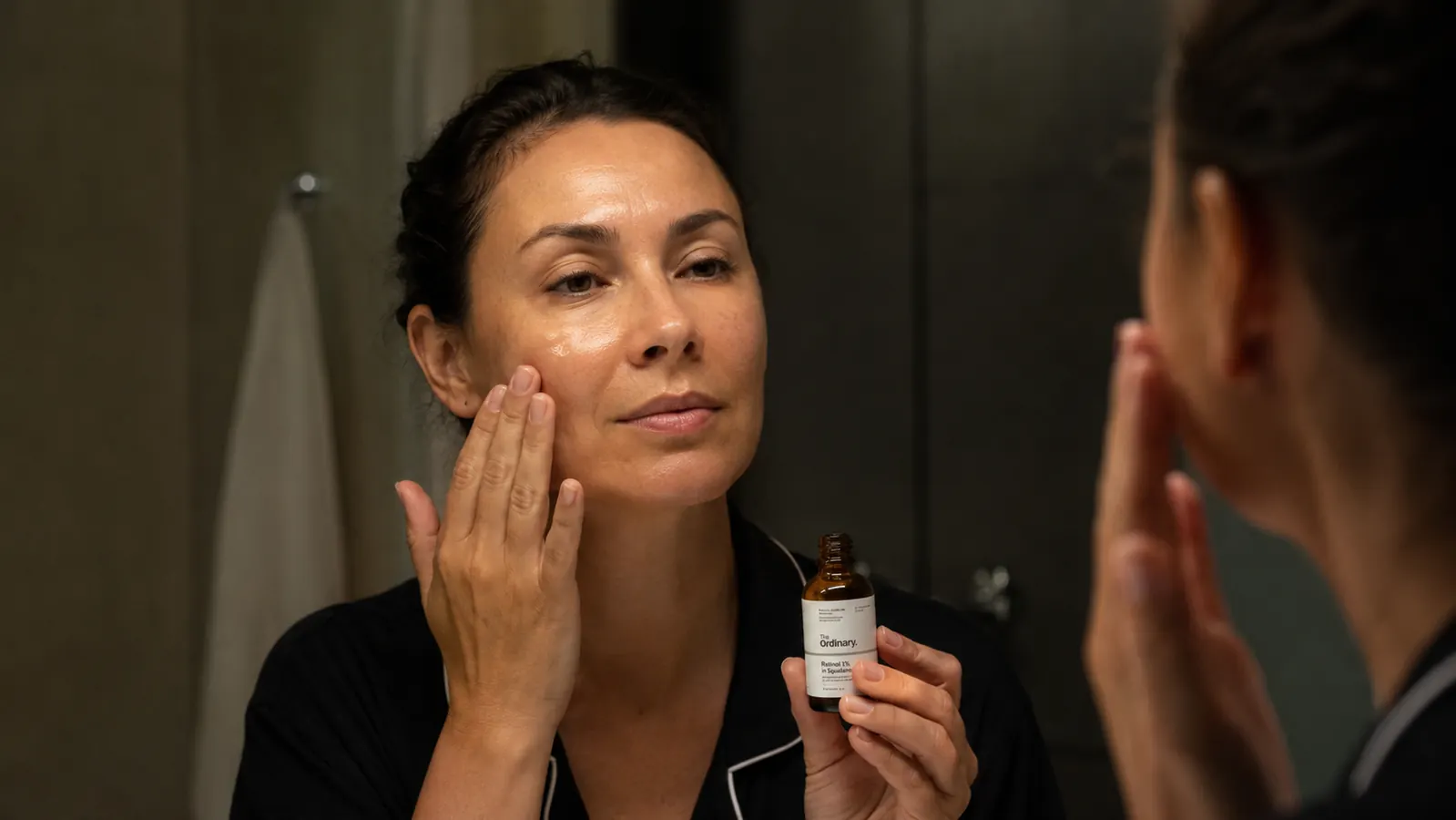 Person applying serum with fingertips to cheek while looking in a well-lit bathroom mirror