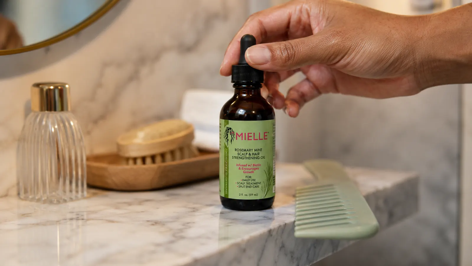 Close-up of the Mielle Organics Rosemary Mint Hair Oil bottle resting on a marble bathroom shelf next to a comb