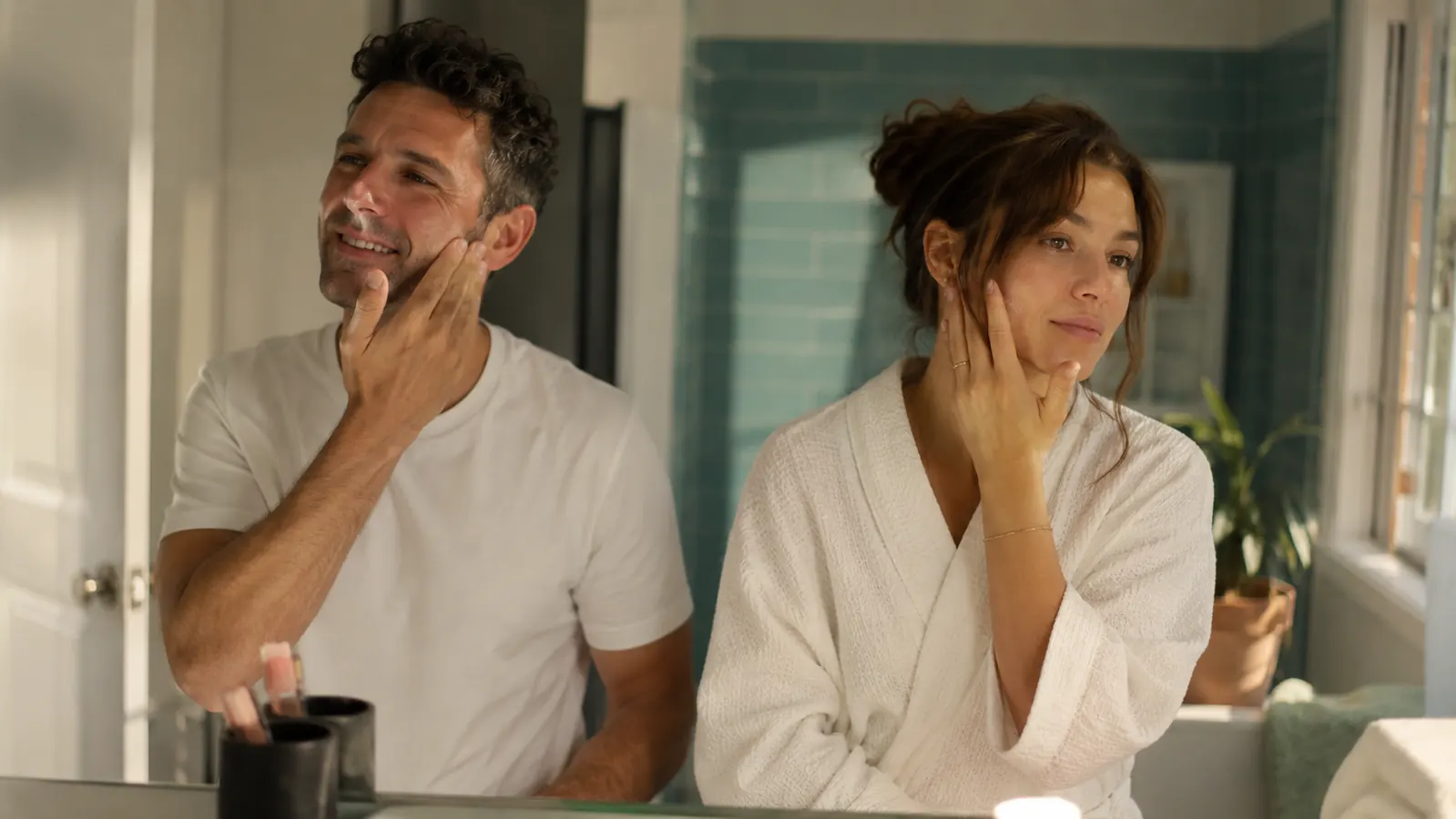 Two people standing at a bathroom mirror, morning light, both with bright and rested-looking skin